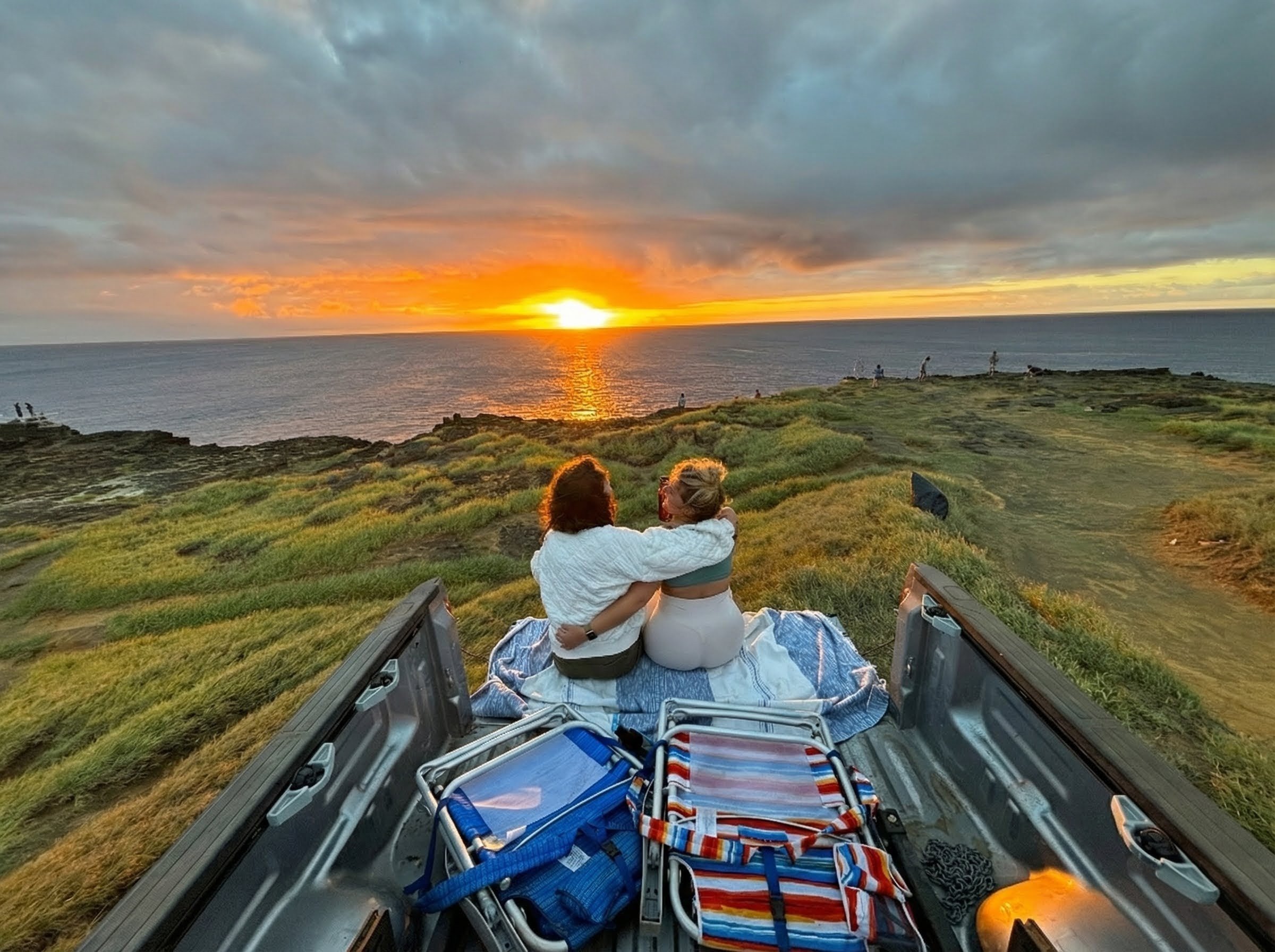 A couple sitting in the back of a jeep watching the sunset over the ocean.